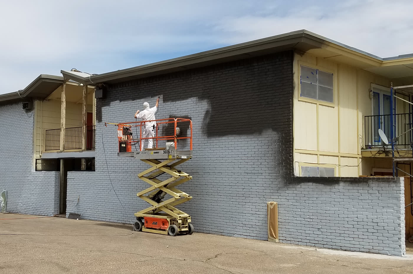 Painter on a scissor lift, halfway through painting a brick building dark grey